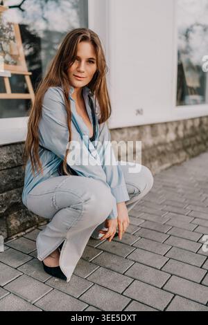 Donna alla moda in denim che posa una giovane donna elegante su asfaltatrici in pietra all'aperto indossando una camicia azzurra e jeans durante una giornata di sole Foto Stock