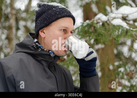 Uomo di mezza età sorseggiando tè caldo da un thermos d'argento in una foresta innevata. Un gentiluomo con una sciarpa bianca e blu a fantasia si gode una pausa con una bevanda calda durante una passeggiata con il freddo tra gli alberi innevati Foto Stock
