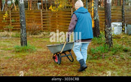Donna anziana che spinge la carriola nel cortile vista da dietro il giorno all'aperto. L'anziano proprietario di casa cammina con la carriola carica attraverso la proprietà all'aperto. I pensionati attivi mantengono il cantiere con una certa stabilità Foto Stock