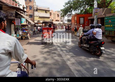 Vivace scena urbana della vecchia Delhi, India, con pendolari che viaggiano in bicicletta in risciò e moto attraverso una stretta strada della città costeggiata da negozi Foto Stock