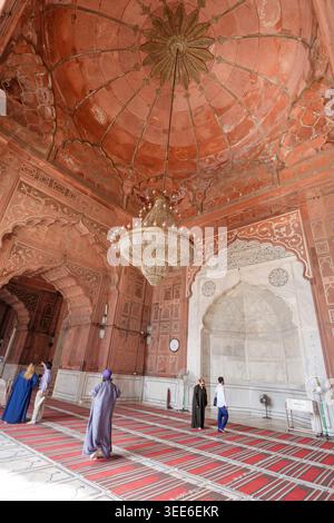 L'interno della grande moschea Jama Masjid a Delhi, India, presenta archi di arenaria rossa e un grande lampadario centrale con i visitatori nella sala di preghiera Foto Stock