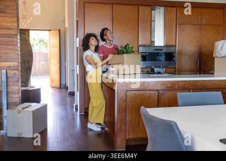 Diverse coppie di donne che disimballano piante e scatole a Kitchen Island, adolescenti che indossano tute, spazio per copiare. Famiglia, lavoro di squadra, luce solare, calore, armadietti, lavorazione del legno, arredamento Foto Stock