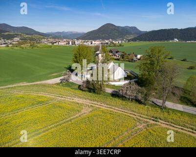 Vista aerea della storica Chiesa di tutti i Santi circondata da un muro di pietra, lussureggianti campi verdi e fiori di colza gialli sotto un cielo azzurro limpido a Lu Foto Stock