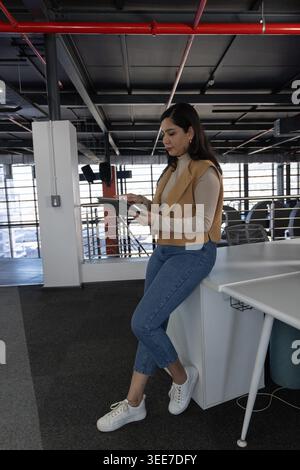 Donna asiatica seduta sul bordo bianco della scrivania utilizzando un tablet nel mezzanino dell'ufficio con gilet beige Foto Stock