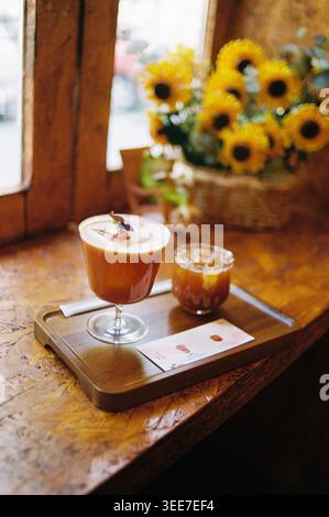 Due bevande artigianali di colore arancione su un vassoio di legno vicino alla finestra di un caffè, decorate con girasoli in un cestino intrecciato, calda e accogliente estetica vintage. Foto Stock