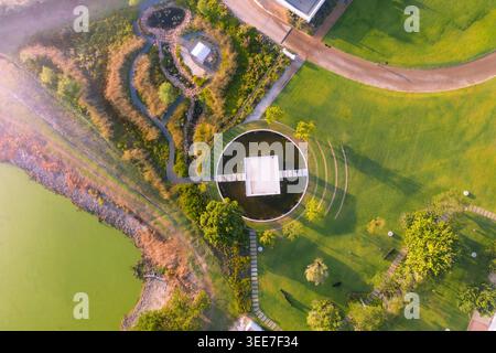 Vista aerea di un moderno giardino paesaggistico caratterizzato da un laghetto circolare con un padiglione centrale, tortuosi sentieri in pietra e lussureggianti prati verdi sotto soffici mattoni Foto Stock