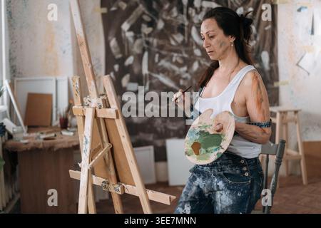donna artistica concentrata che tiene la tavolozza davanti al cavalletto utilizzando tela con immagine, pittura con pittura ad olio. copia spazio Foto Stock