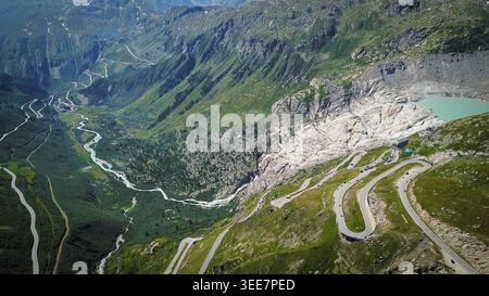 Vista aerea del passo Furka con i suoi tornanti iconici, il ghiacciaio del Rodano e un lago turchese di montagna annidato nelle Alpi svizzere vicino a Oberg Foto Stock