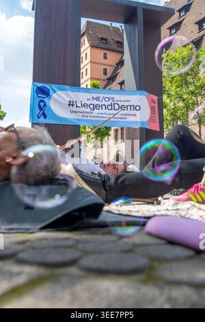 9 maggio 2026, Baviera, Norimberga: Le bolle di sapone volano davanti a persone sdraiate sul pavimento mentre uno striscione chiede una "demo sdraiata". In occasione della "giornata internazionale di sensibilizzazione ME/CFS”, i malati, i parenti e i sostenitori richiamano l'attenzione sulla situazione delle persone affette da ME/CFS, una grave malattia multisistemica che finora è stata poco studiata. Circa 400 persone partecipano alla manifestazione a Norimberga. Foto: Pia Bayer/dpa Foto Stock