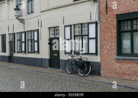 Bicicletta con piedistalli accanto alle facciate di case in mattoni bianchi sulla strada acciottolata di Bruges, Belgio, finestre con persiane, atmosfera urbana silenziosa. Ciclismo Foto Stock