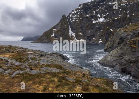 Le onde si schiantano contro le scogliere rocciose del Lofoten mentre le nuvole scure si insinuano in alto. Le cime innevate si stagliano maestosamente, mostrando la selvaggia e selvaggia Norvegia Foto Stock