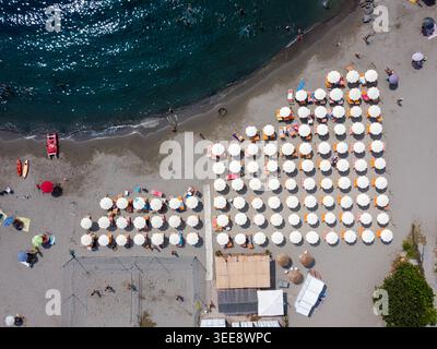 Vista aerea di file organizzate di ombrelloni bianchi e lettini sulla spiaggia sabbiosa con persone che nuotano nel mare cristallino di San Lorenzo, Liguria Foto Stock