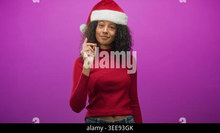 Adolescente con il cappello di babbo natale che tiene un piccolo oggetto con la mano vicino al viso mentre sorride in studio su uno sfondo viola; gioia per le vacanze. Foto Stock