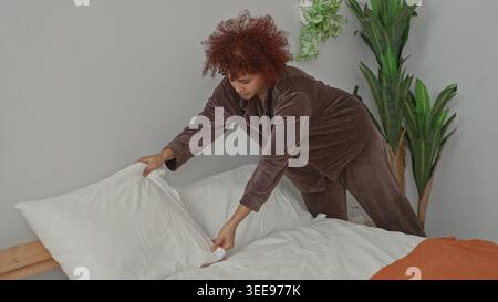 La donna mette il cuscino sul letto e leviga le lenzuola bianche mentre nasconde un tiro arancione accanto a una testata di legno in una camera da letto dell'edificio; calma routine. Foto Stock