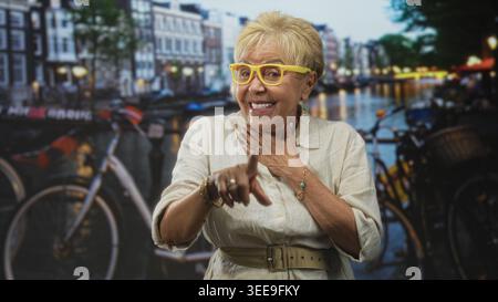 Donna anziana che punta il dito con la mano sul petto in uno studio con bici e canale sullo sfondo e occhiali gialli; calore gioioso. Foto Stock