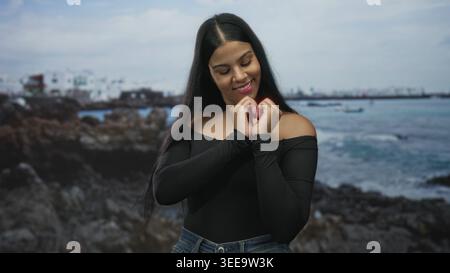 Donna che forma un gesto cardiaco con le mani sulla costa rocciosa sotto il cielo nuvoloso del mare; amore affetto gioia romanticismo. Foto Stock