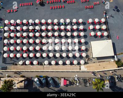 Vista aerea di file di ombrelloni bianchi e lettini rossi su una spiaggia di ciottoli scuri con persone che camminano e parcheggiano auto lungo il lungomare di Imperia, Ligu Foto Stock