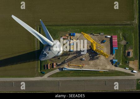 Vista aerea di una turbina eolica in costruzione con una grande gru gialla, pale della turbina a terra e contenitori di spedizione in un campo verde. Foto Stock