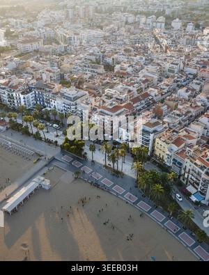 Vista aerea della città costiera di Sitges con la sua spiaggia sabbiosa, la passeggiata costeggiata da palme e la densa architettura urbana sotto la calda luce del sole a Sitges, Cata Foto Stock