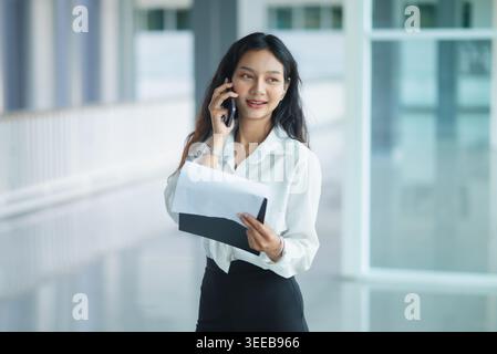 Una donna d'affari sorridente parla su uno smartphone mentre tiene una cartella in un ufficio moderno e luminoso, mostrando la comunicazione con il cliente, il coordinamento aziendale, il papa' Foto Stock