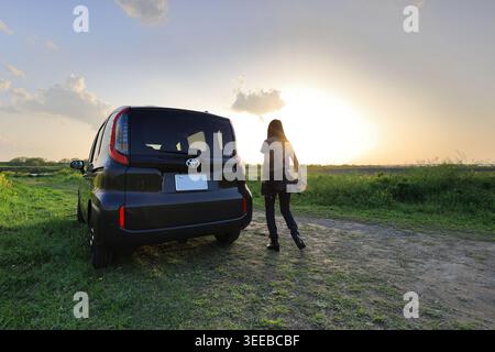 Una giovane donna si trova accanto alla sua auto compatta in un campo aperto, catturando una foto del tramonto dorato mozzafiato che dipinge il cielo ampio. Foto Stock