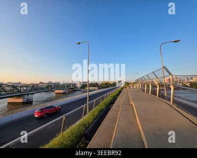 Tranquillo paesaggio mattutino della città caratterizzato da un passaggio pedonale vuoto lungo la strada asfaltata con un ponte di acciaio rosso che attraversa il fiume sotto la splendida cl Foto Stock