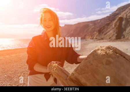 Donna in spiaggia sorridente alla luce del sole mentre tocca un vecchio pezzo di legno su una riva costiera Foto Stock