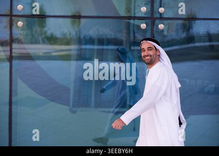 L'uomo sorridente degli Emirati che indossa una kandura bianca e un ghutra cammina con sicurezza accanto a una moderna facciata in vetro, il suo riflesso visibile. Ritratto di stile di vita che suggerisce cultura, professionalità, ottimismo e vita urbana contemporanea. Foto Stock