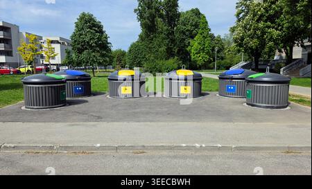 Sei contenitori circolari semi-sotterranei con lati grigi a strisce e vivaci coperchi codificati a colori si trovano su un'area pavimentata vicino a un parco verde e residente Foto Stock