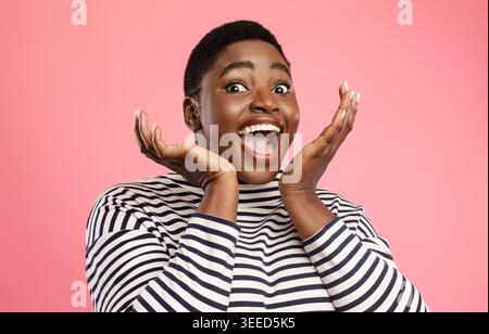 Donna Nera gioiosa che grida tenendosi le mani vicino al viso, sfondo rosa Foto Stock