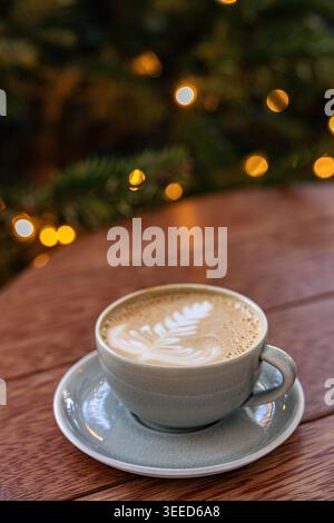 Tazza da caffè latte con latte art, su un tavolo di legno, caratterizzato da un albero di Natale leggermente sfocato con calde luci fiabesche sullo sfondo. Foto Stock
