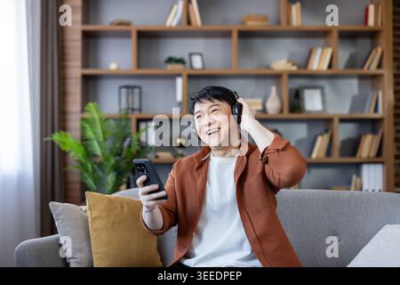 L'uomo asiatico è felice di ascoltare la musica sul suo smartphone con le cuffie wireless mentre è seduto comodamente su un divano in un moderno salotto di casa, godendo del tempo libero Foto Stock