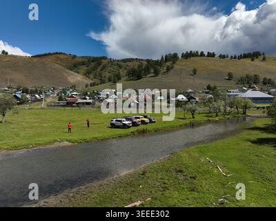 Due persone si trovano su un terreno erboso accanto a un fiume tranquillo con diversi veicoli parcheggiati nelle vicinanze, che si affacciano su un villaggio rurale Foto Stock