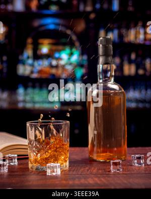 Il whisky dinamico si tuffa in un bicchiere di cristallo con bottiglia, cubetti di ghiaccio e un libro aperto su un caldo pub bar irlandese di notte. Foto Stock