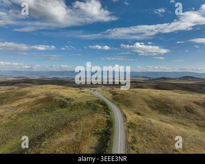 Una prospettiva aerea cattura una lunga strada asfaltata curvilinea che taglia le colline aperte e ondulate ricoperte da una vegetazione secca dorata-marrone sotto un ampio cle Foto Stock