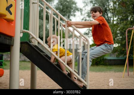 Due fratelli che condividono il divertimento giocoso sullo scivolo del parco giochi insieme. Momento di legame tra fratelli e sorelle mentre i bambini più grandi e i più piccoli amano le attività all'aperto. Interazione tra fratelli e sorelle con le colorate attrezzature da gioco nel parco Foto Stock