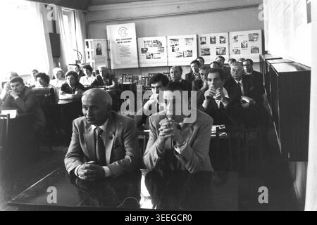 Slaviansk, 1983. Sala conferenze della fabbrica di isolatori per armature (AIZ). Due uomini in tuta siedono in prima fila del pubblico, ascoltando da vicino una presentazione durante una riunione ufficiale della fabbrica. Questa fotografia vintage in bianco e nero cattura l'atmosfera autentica dell'epoca, riflettendo le attività quotidiane e la cultura industriale della regione durante il periodo sovietico. Funge da prezioso documento storico della comunità locale, preservando la memoria visiva delle persone e della loro vita quotidiana, mostrando un tranquillo Donbas. Foto Stock