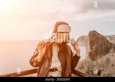 Donna viaggiatrice sulla costa in piedi sul bordo di una scogliera che indossa un caldo cappello invernale e una giacca di pelle durante il tramonto dell'ora d'oro Foto Stock