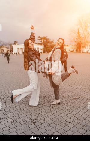 Donne che ballano amicizia due amici eleganti si tengono per mano e ballano gioiosamente su una piazza acciottolata durante un caldo tramonto Foto Stock