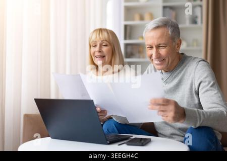 Felice coppia matura seduta insieme a casa, sorridendo mentre rivede i documenti cartacei Foto Stock
