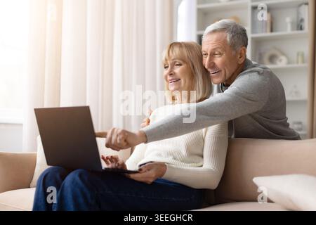 Allegra coppia matura seduta insieme a casa, uomo che punta sullo schermo del notebook Foto Stock