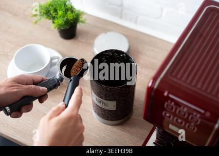 Primo piano di mani che riempiono il portafiltro con caffè macinato accanto a una macchina da caffè rossa e al contenitore del caffè su un piano di lavoro della cucina. Caffè a casa pr Foto Stock
