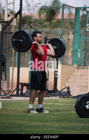 L'uomo arabo mediorientale esegue uno squat frontale CrossFit con barbell in una palestra all'aperto, concentrandosi sull'allenamento di forza e fitness mentre si fa esercizio con un amico in un'area di allenamento all'aperto. Foto Stock