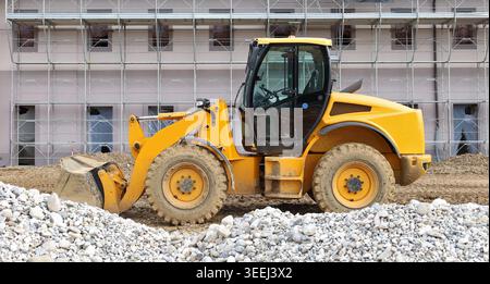 Pala caricatrice gommata in un'area sporca con un mucchio di ghiaia davanti e un edificio in costruzione dietro. Vista laterale Foto Stock