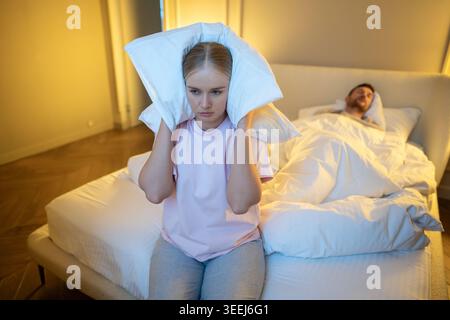 Moglie irritata seduta sul letto che copriva le orecchie con un cuscino per bloccare il suono ruvido mentre l'uomo dorme. Foto Stock