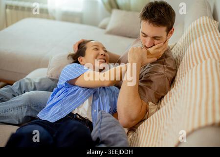 Una donna coreana siede vicino a un uomo caucasico in un soggiorno. Stanno ridendo e si godono il loro tempo insieme su un divano durante il pomeriggio. Foto Stock