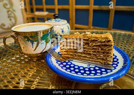 Una fetta di torta al miele a strati su un piatto di ceramica blu con una forchetta, servita con una tazza di caffè cappuccino in un accogliente caffè. Foto Stock