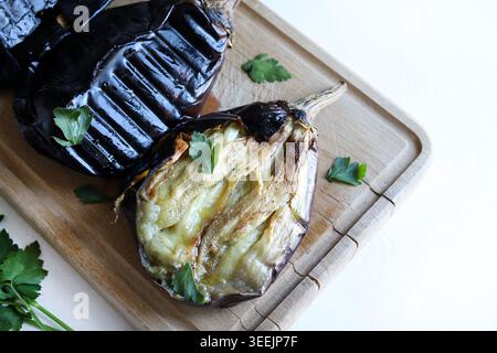 Melanzane arrostite e una parzialmente sbucciata su un tagliere di legno con prezzemolo. Vista dall'alto dell'ingrediente alimentare sano. Cucina casalinga. Vista dall'alto. Foto Stock
