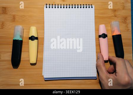 Flat lay di quaderno a griglia aperto con pennarelli colorati. Mano che tiene un evidenziatore rosa su sfondo di legno. Vista dall'alto delle forniture per ufficio per Foto Stock