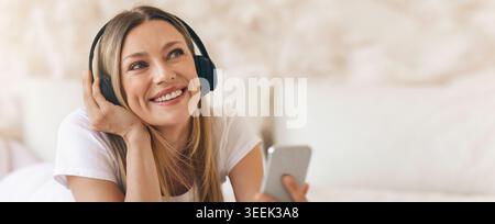 La donna gode di musica mentre è sdraiata sul letto in una camera accogliente con smartphone e cuffie Foto Stock